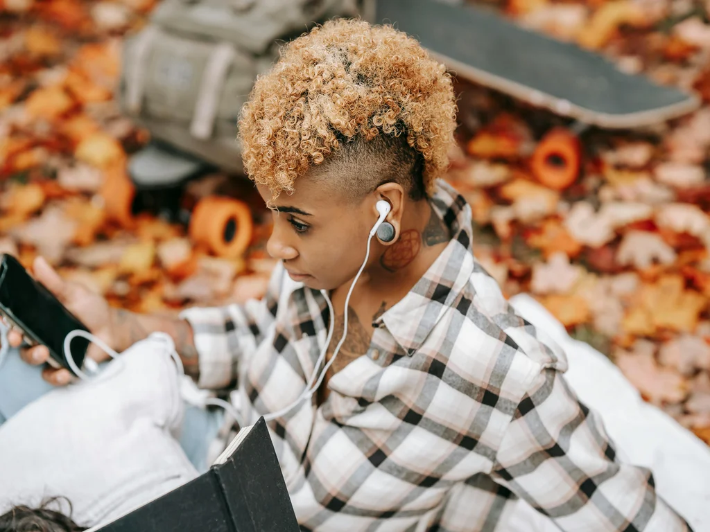 jeune femme noire qui porte des écouteurs filaires dans les oreilles. De profil gauche, elle est assise et porte une chemise à carreaux blanche et noire