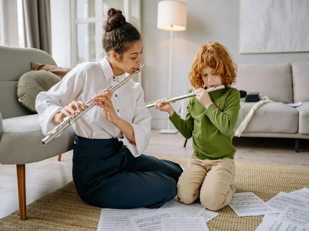 une jeune femme blanche joue de la flute à côté d'un enfant blanc et roux qui joue aussi de la flute. Des partitions sont étalées par terre devant eux. 